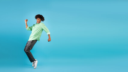Fototapeta premium Excited african american teen guy fooling, standing on sneakers tiptoes, dancing and having fun over blue background, panorama with free space, studio shot