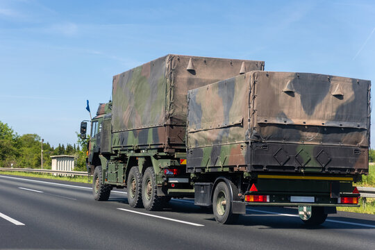 Europe military truck camouflage drives along autobahn green forest landscape during summer daylight. Tactical transport vehicle logistic defense mobility convoy movement European road infrastructure