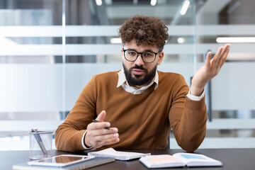 Young businessman actively communicating and discussing concepts during an online video call from his modern office workspace, wearing glasses and a sweater