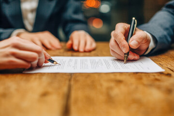 Two businesspeople in formal suits signing a legal contract paper together on a wooden table in a professional setting with warm lighting and blurred background ligh