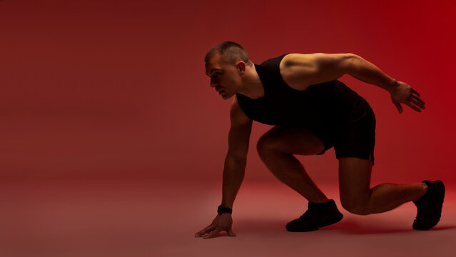 Side view of sportsman standing on start, male sprinter in sportswear ready to run race or marathon, red background, panorama with free space