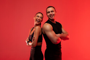 Happy sporty couple standing back to back and smiling, man and woman wearing black workout gear, posing over red neon studio background