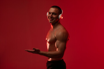 Man with muscular body wearing wireless headphones and pointing aside, holding hand with open palm against red studio background