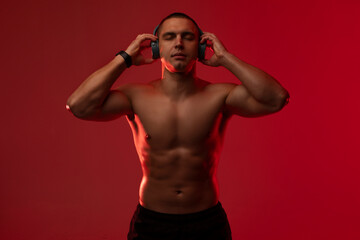 Fototapeta premium Male athlete with bare torso listening to music in wireless headphones with closed eyes, standing over red studio background. Sport and technology concept