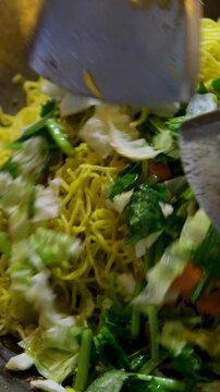 stir with large spoons in a wok topview a plate of stir fried yellow noodle with lots of saag veges.