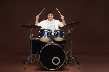 Smiling teenage boy with drumsticks learning how to play drums on brown background