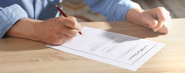 Senior man signing Last Will and Testament at wooden desk indoors, closeup