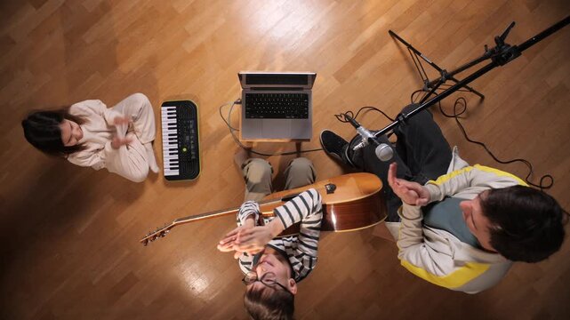 Family members, including a young girl, boy, and adult, sitting on a wooden floor, learning and playing musical instruments with a laptop and microphone setup for online education