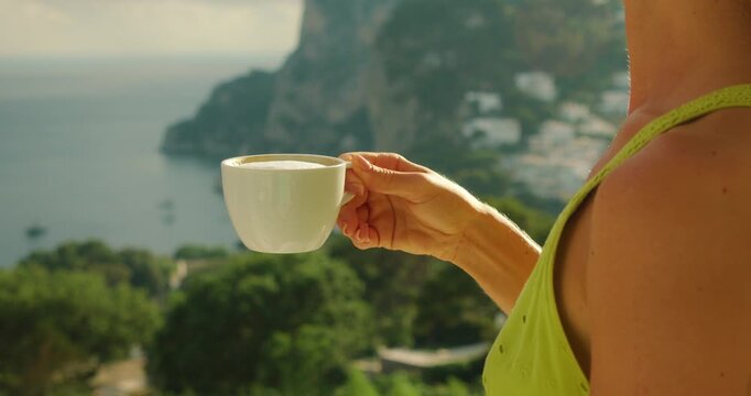 A woman enjoys a morning coffee while overlooking the coastline and sea on a summer day in Capri Italy