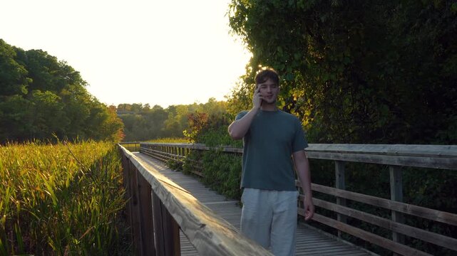 Young man walks toward and past camera while speaking on phone along wooden boardwalk. Royal Botanical Garden, Burlington, Ontario.