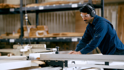 Male carpenter cutting wooden plank with a table saw, wearing safety glasses and ear protection in a woodworking factory