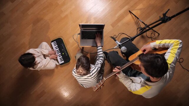 Young artists collaborating on a music project, learning sound production, recording, and musical arrangement using a guitar, keyboard, microphone, and laptop in a home studio setup