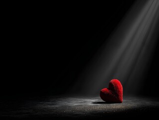 Small red velvet heart resting on a dark surface with dramatic spotlight beams and wide copy space.