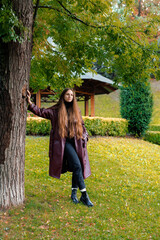 Woman in long coat leans against tree in park with green foliage