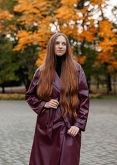 Woman in burgundy coat stands outdoors among autumn foliage