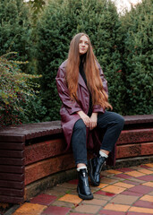 Young woman in long coat sitting on bench in park surrounded by greenery