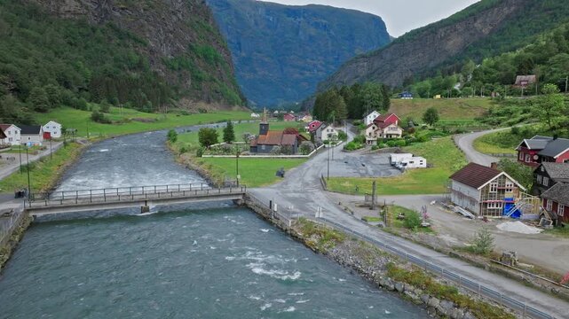 Aerial view of bridge and river flowing through Flam village in Norway. Drone footage shows roads, houses and green valley landscape surrounded by steep mountains.