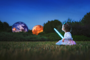 Little child with a glowing light sword looking at large illuminated planet models in a night park. Concept of space dreams and imagination.