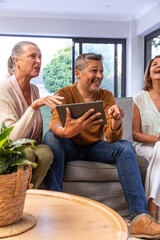 Senior woman with two mature friends sitting in living room, sharing tablet on gray sofa