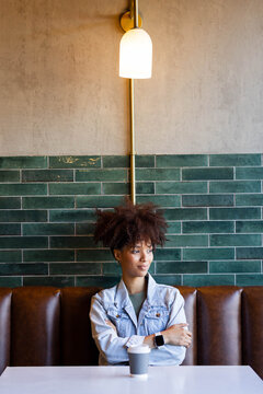 African American woman sitting in cafe booth at white table wearing denim jacket holding to-go cup