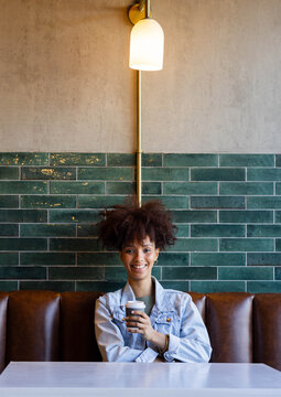 African American adult woman smiling, holding takeout cup, wearing denim jacket by green tile wall