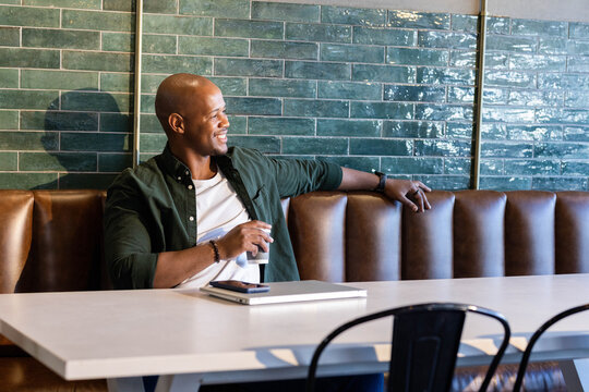 Man leaning back in brown leather booth at cafe, holding takeout cup by closed-laptop stacked phone