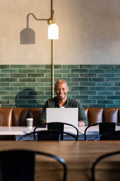 Adult African American man sitting at long cafe table working on silver laptop wearing green shirt