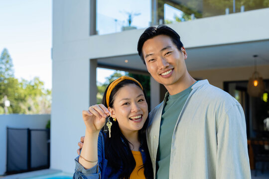 Asian couple smiling, woman holding house keys, wearing yellow top at two-story house terrace
