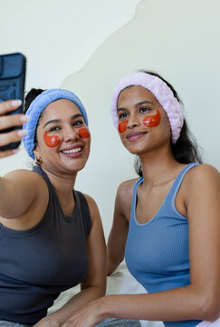 Diverse female friends sitting on bed taking selfie with phone headbands under-eye patches