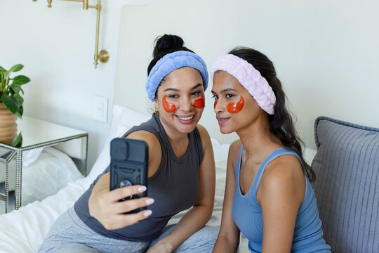 Diverse female friends taking selfie with phone case on bed wearing headbands and eye patches