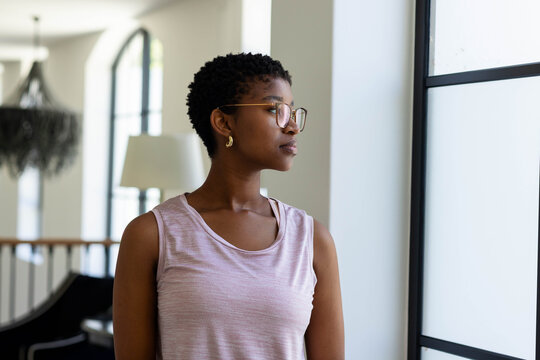 African American woman standing, gazing out multi-pane window in living room wearing sleeveless top