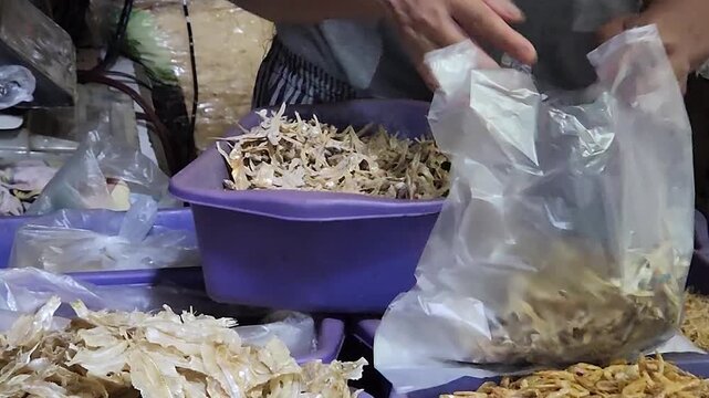 Dried fish vendor scooping seafood into plastic bag at market stall. Hands packing salted fish strips for retail sale in traditional market. Street market seafood seller bagging dried fish ingredients