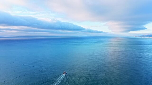A small boat moves across the vast ocean water during early morning hours. The sky is filled with clouds reflecting light over the blue water. The scenery is peaceful and natural.