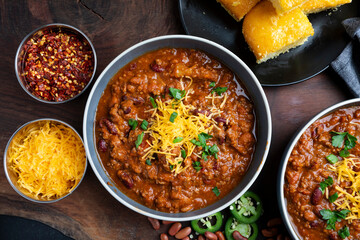 A rustic overhead view of homemade beef chili served in bowls and topped with shredded cheddar cheese, fresh herbs, and sliced jalape&ntilde;os. Surrounded by red beans, chili flakes, and cornbread on a wood