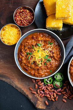 beef chili served in bowls and topped with shredded cheddar cheese, fresh herbs, and sliced jalape&ntilde;os. Surrounded by red beans, chili flakes, and cornbread on a wooden table, this image conveys comfor