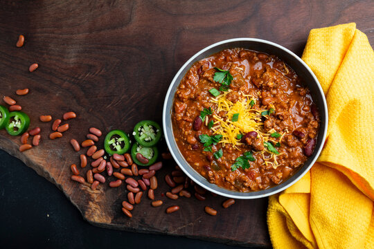 A rustic overhead view of homemade beef chili served in bowls and topped with shredded cheddar cheese, fresh herbs, and sliced jalape&ntilde;os. Surrounded by red beans, chili flakes, and cornbread on a wood