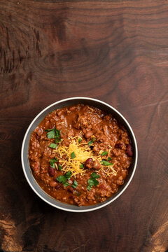 A rustic overhead view of homemade beef chili served in bowls and topped with shredded cheddar cheese, fresh herbs, and sliced jalape&ntilde;os. Surrounded by red beans, chili flakes, and cornbread on a wood