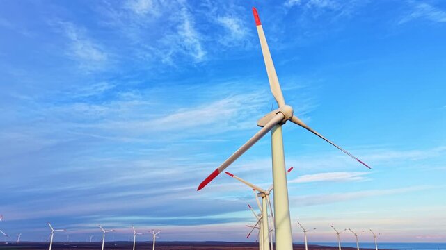 Rows of large wind turbines stand tall against a bright sky while producing renewable energy. The scene is set near a body of water in an open area with a flat landscape.