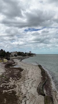 Aerial drone flying forward along the shoreline in Puerto Progreso, revealing a quiet beach, coastal buildings, and calm sea under an overcast sky, captured in D-Log.