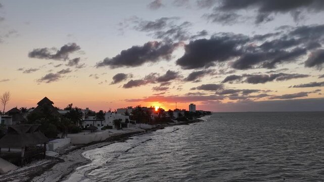 Aerial sunset view in Puerto Progreso, Yucat&aacute;n, with the drone hovering over the sea as the sun sets on the horizon, dramatic clouds, calm waves, and a quiet coastal shoreline.