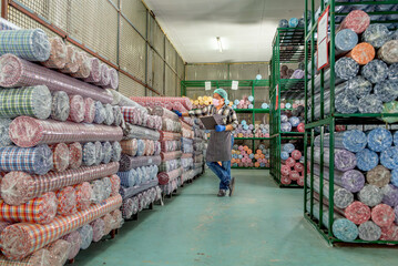 Warehouse worker in mask and gloves standing with pallet Jack holding clipboard and checking inventory of colorful fabric rolls on shelves. Male staff auditing textile stock in storage warehouse.