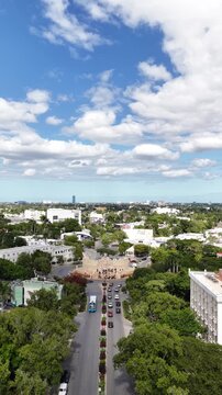 Vertical aerial view in M&eacute;rida, Yucat&aacute;n, as the drone flies forward over Paseo Montejo toward the Altar a la Patria roundabout, with lush trees, city traffic, and a blue, cloud-filled sky.