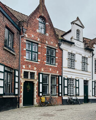 Naklejka premium Bruges, Belgium – December 27, 2025. Traditional brick houses with white facades, balconies and green shutters on a quiet cobblestone street in the historic old town.