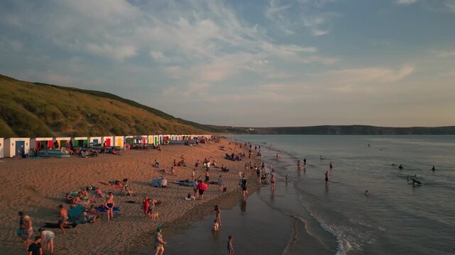 A vibrant beach scene at dusk with people relaxing, playing, and surfing. Rows of colorful beach huts line the sand.