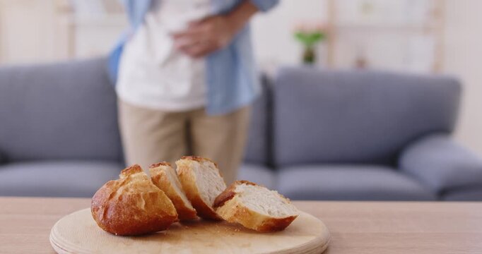 Gluten bread trigger of food intolerance and allergy on wooden board, stomach pain and cramps, indigestion of woman standing nearby. Person holding belly by living room sofa, suffering from celiac gut