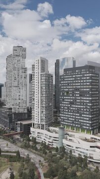 Aerial drone flying backwards from Santa Fe skyscrapers revealing Parque La Mexicana with cloudy blue sky in M&eacute;xico City. Vertical video shot in D-Log M