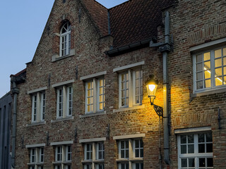 Naklejka premium Bruges, Belgium – December 27, 2025. Historic brick house facade with traditional Flemish gable, warm street lantern and illuminated windows at blue hour, atmospheric European old town scene.
