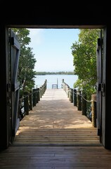 Fototapeta premium A wooden dock extends toward calm water, framed by open barn-style doors and lush green mangroves near Mary Selby Gardens Historic Spanish Point, Sarasota, Florida, U.S