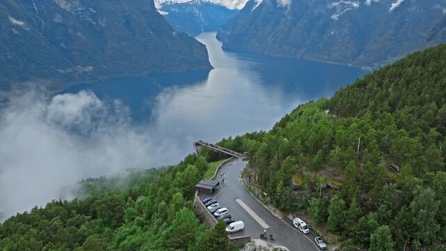 High aerial view of Stegastein viewpoint and parked cars above Aurlandsfjord. Norwegian mountain road and platform overlook deep fjord scenery and green slopes.