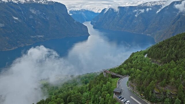 Drone shot of Stegastein road and viewpoint above Aurlandsfjord, Norway. Winding road and forested cliffs appear above clouds drifting across the fjord.